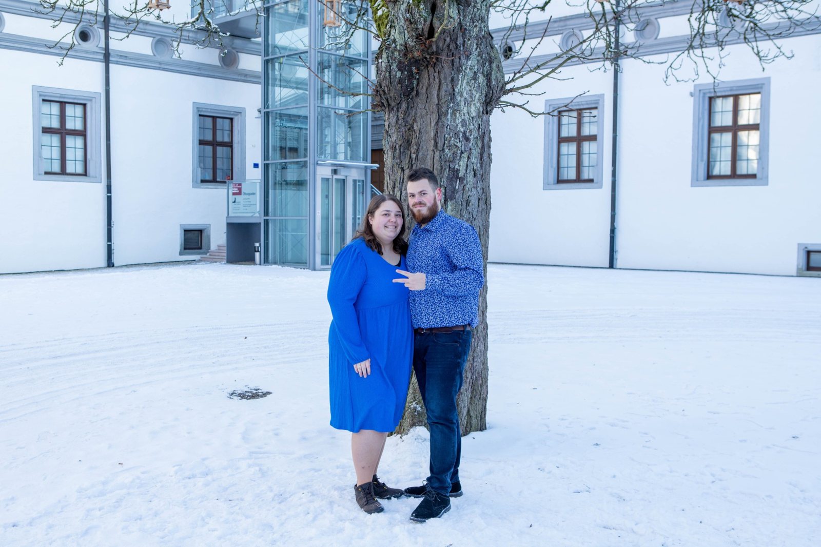 Paar im Schnee vor einem Baum, der Mann zeigt lachend ein Peace-Zeichen
